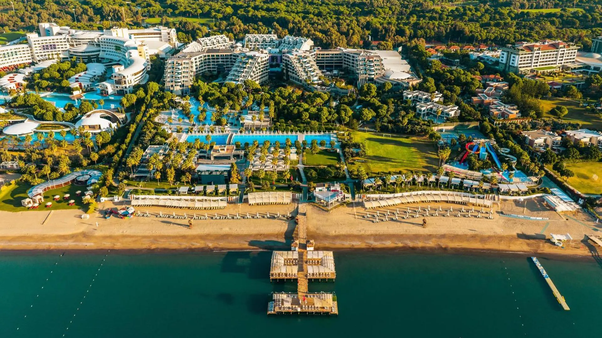 Susesi Luxury Resort Belek Exterior photo This is an aerial photograph showcasing a large, sprawling resort complex set against a backdrop of lush greenery, bordering a body of water.

**Key features in the photo include:**

*   **The Resort:** A vast complex of white, multi-story buildings 